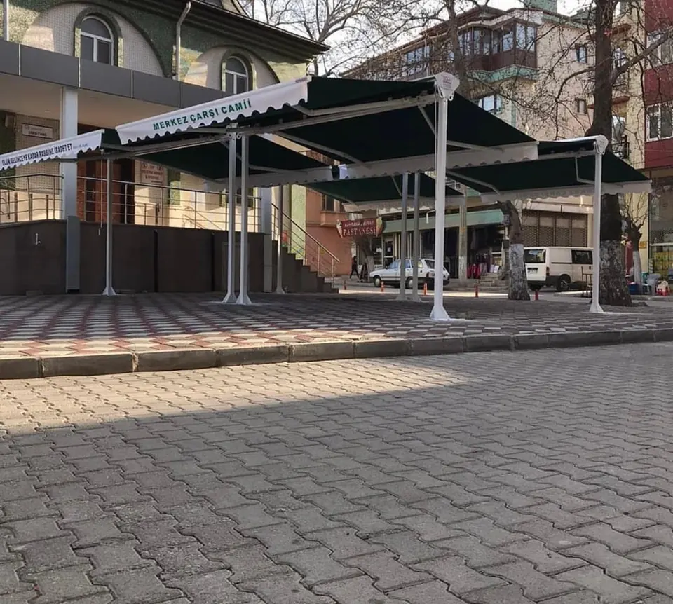 Cami avlusuna kurulan, saçağı baskılı yeşil çift açılır tente sistemi ve beyaz taşıyıcı dikmeler