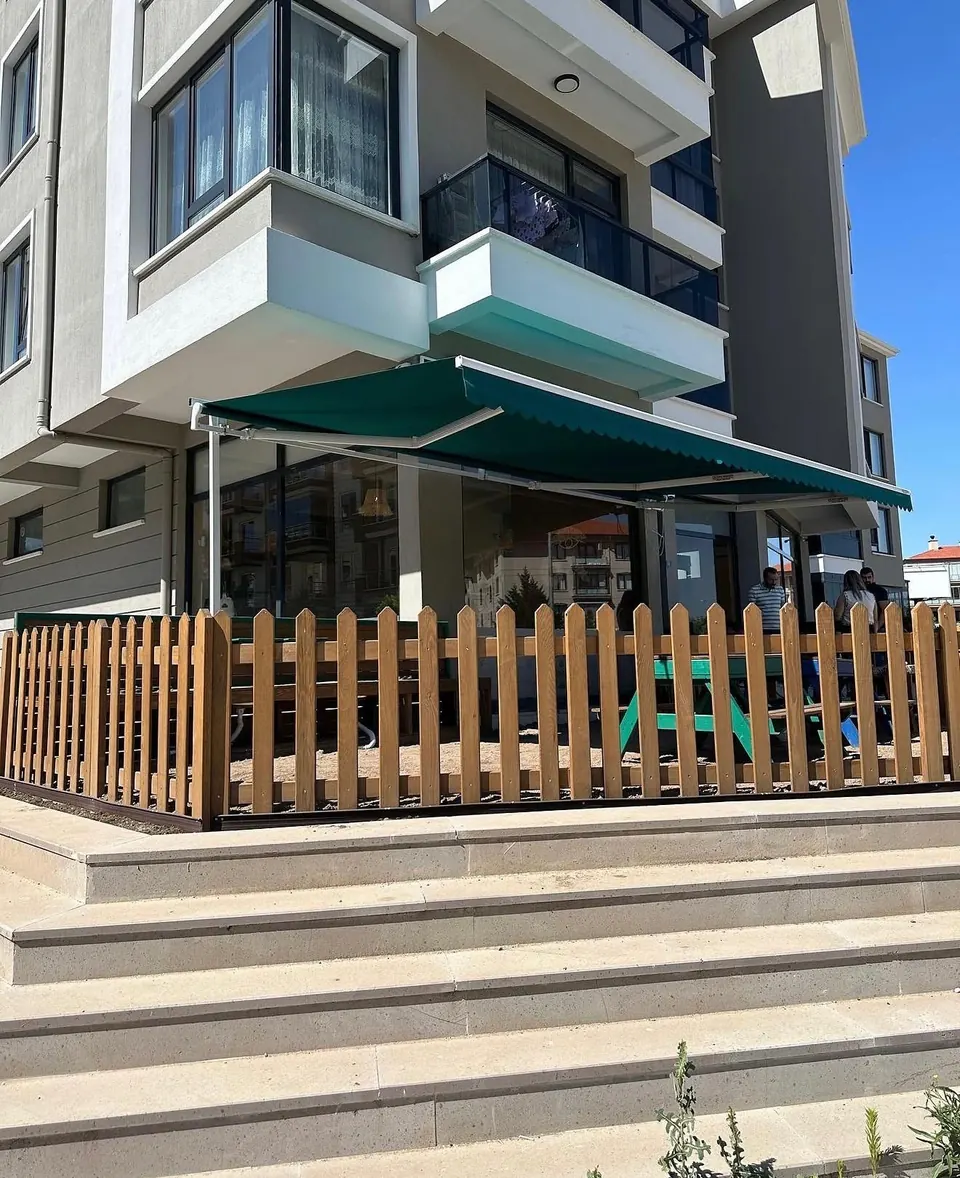 Apartman zemin katındaki kafe bahçesini gölgeleyen geniş yeşil mafsallı tente