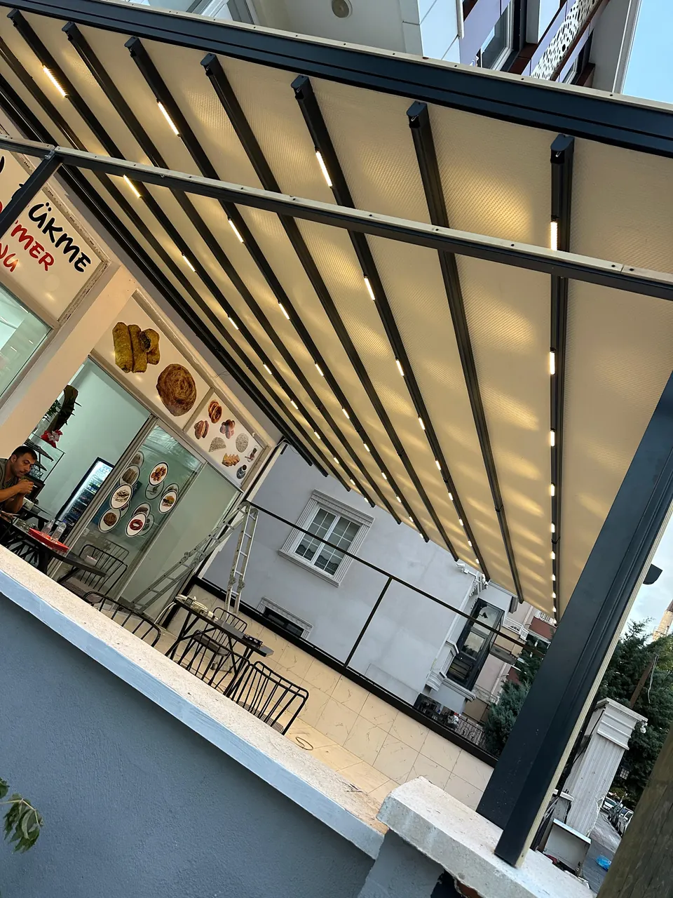 Kafe terasında LED aydınlatmalı, raylı açılır-kapanır pergole tente sisteminin içeriden görünümü